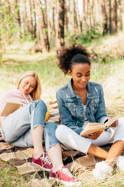 Два багатоетнічні друзі сидять на плетеній ковдрі і читають книги — стокове фото