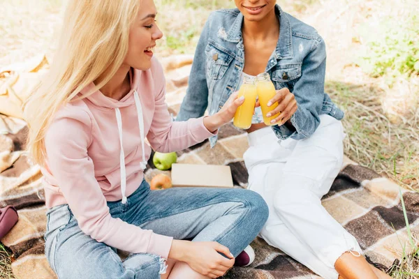 Cropped view of two multicultural friends sitting on plaid blanket with bottles of orange juice — Stock Photo