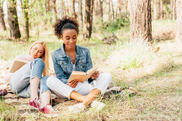 Два багатоетнічні друзі сидять на плетеній ковдрі і читають книги — стокове фото