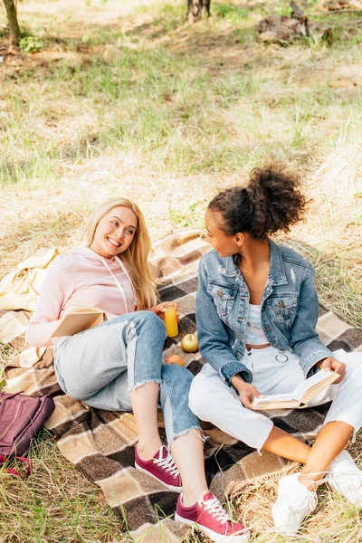 Deux amis multiethniques assis sur une couverture à carreaux et lisant des livres — Photo de stock