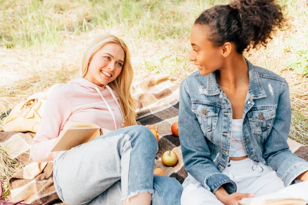 Due amici multietnici seduti su una coperta a quadri e leggendo libri — Foto stock