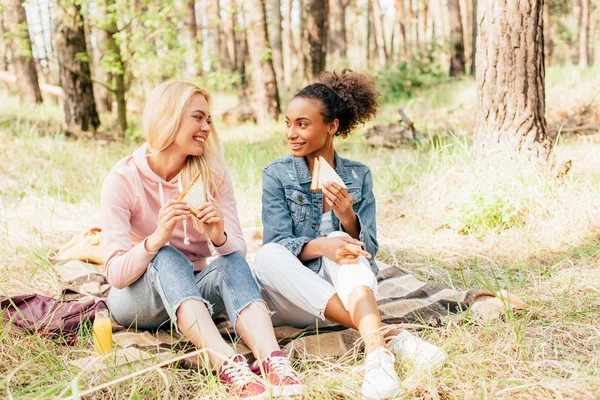 Two smiling multiethnic friends sitting on plaid blanket with sandwiches and looking at each other — Stock Photo
