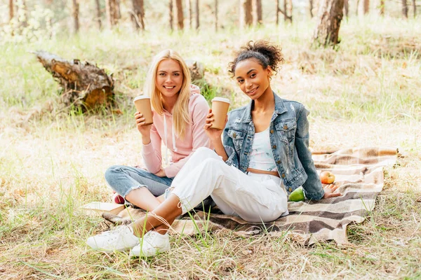 Two multiethnic friends sitting on plaid blanket and holding paper cups of coffee — Stock Photo