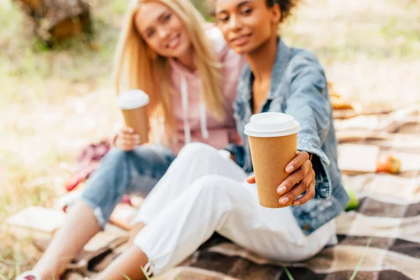 Vista cortada de dois amigos multiétnicos sentados no cobertor xadrez e segurando xícaras de papel de café — Fotografia de Stock
