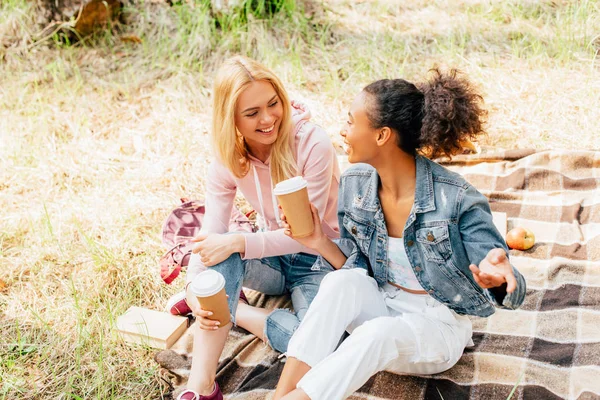 Zwei multiethnische Freunde sitzen auf karierter Decke und halten Pappbecher mit Kaffee in der Hand — Stockfoto