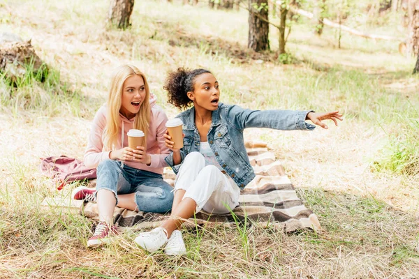 Zwei verblüffte multiethnische Freunde, die auf Plaid mit Pappbechern Kaffee sitzen und wegschauen — Stockfoto