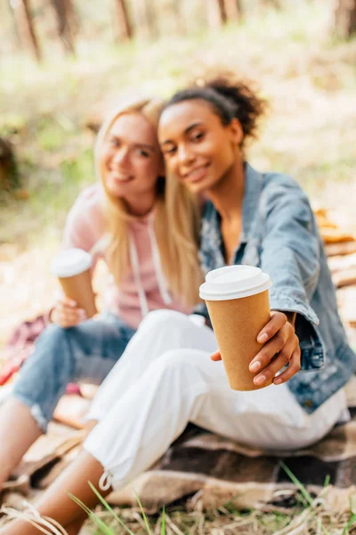 Selektiver Fokus zweier multiethnischer Freunde, die auf karierter Decke sitzen und Pappbecher mit Kaffee in der Hand halten — Stockfoto