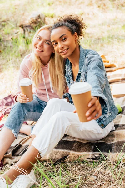 Zwei multiethnische Freunde sitzen auf karierter Decke und halten Pappbecher mit Kaffee in der Hand — Stockfoto