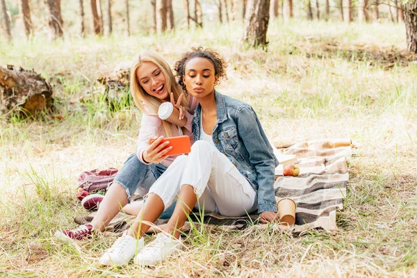 Deux amis multiculturels joyeux avec des tasses en papier de prise de café selfie dans la forêt — Photo de stock