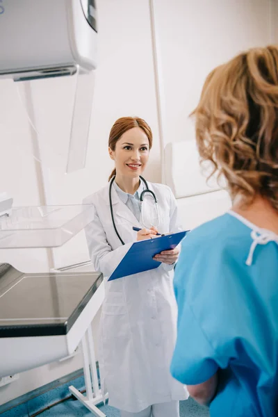 Selective focus of smiling radiographer writing on clipboard near patient in clinic — Stock Photo