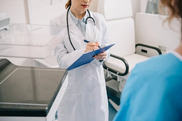 Vista recortada del médico escribiendo en el portapapeles mientras está de pie cerca del paciente - foto de stock