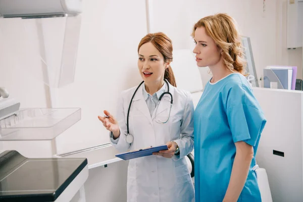 Beautiful young radiologist in white coat showing x-ray machine to patient — Stock Photo