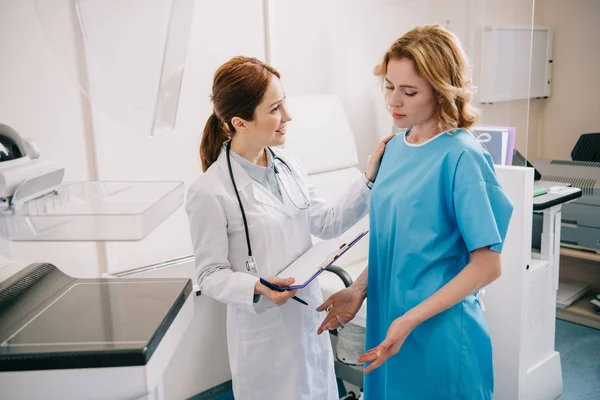 Smiling doctor holding clipboard and touching shoulder of disappointed woman — Stock Photo