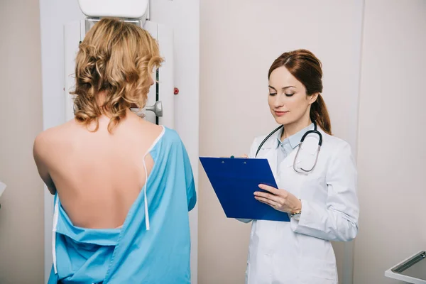 Hermoso médico joven mirando el portapapeles mientras está de pie cerca del paciente durante la prueba de mamografía en la máquina de rayos X - foto de stock