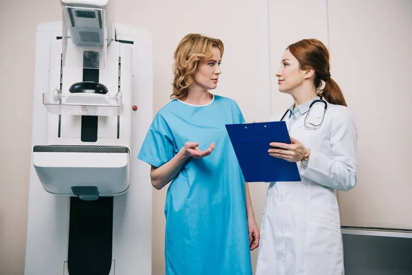 Hermosa mujer hablando con el radiólogo mientras está de pie cerca de la máquina de rayos X - foto de stock