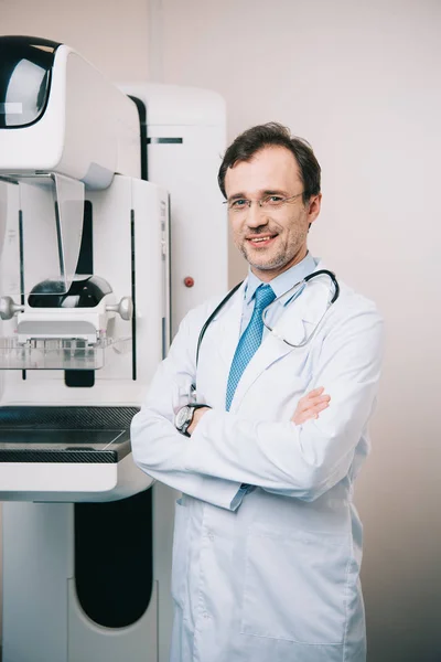 Radiologista sorridente em pé perto da máquina de raios X com braços cruzados e olhando para a câmera — Fotografia de Stock