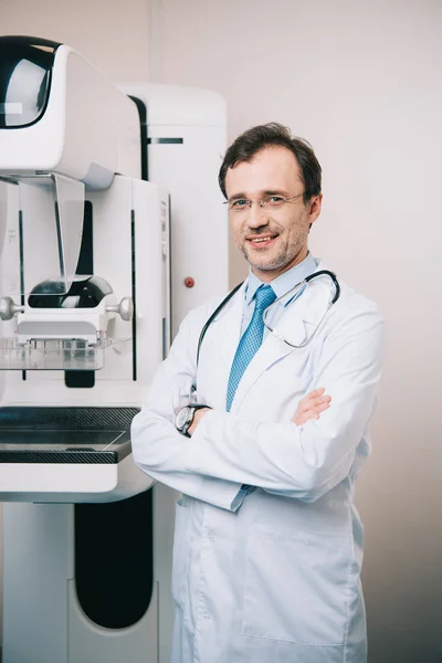 Radiólogo sonriente de pie cerca de la máquina de rayos X con los brazos cruzados y mirando a la cámara - foto de stock