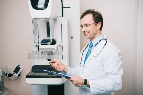 Smiling radiographer standing near x-ray machine and holding clipboard — Stock Photo