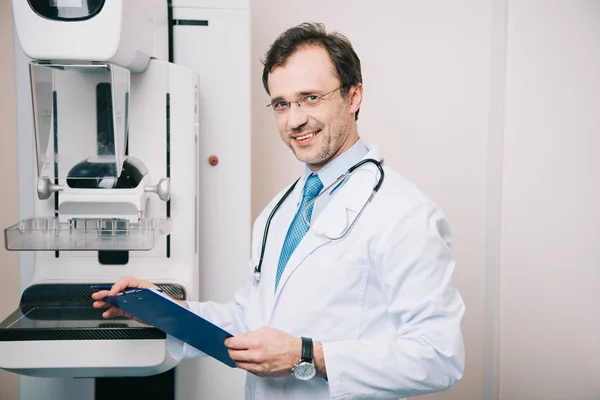 Radiólogo sonriente sosteniendo el portapapeles mientras está de pie en la máquina de rayos X y mirando a la cámara - foto de stock