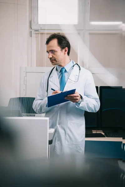 Selective focus of handsome doctor in white coat writing on clipboard — Stock Photo