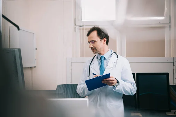 Selective focus of smiling doctor writing on clipboard and looking away — Stock Photo