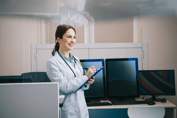 Pretty young doctor in white coat smiling while writing on clipboard — Stock Photo