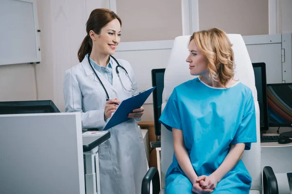 Alegre médico sosteniendo portapapeles cerca hermosa mujer sentado en sillón en el hospital - foto de stock