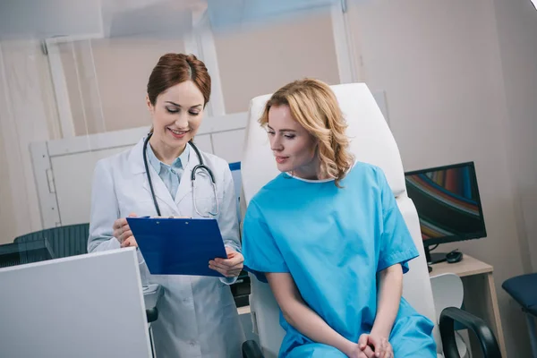 Bastante joven médico y paciente sonriente mirando portapapeles con diagnóstico - foto de stock