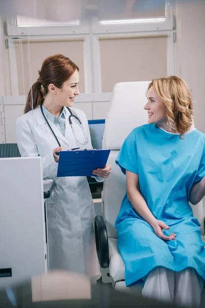 Alegre médico sosteniendo portapapeles mientras está de pie cerca del paciente sentado en el sillón - foto de stock