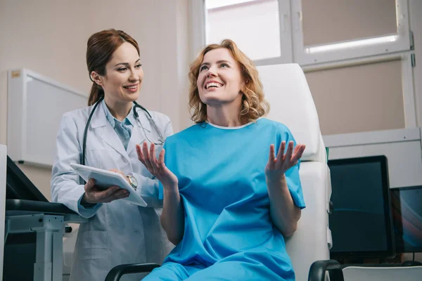 Femme heureuse gestuelle et regardant vers le haut près médecin souriant tenant tablette numérique — Photo de stock