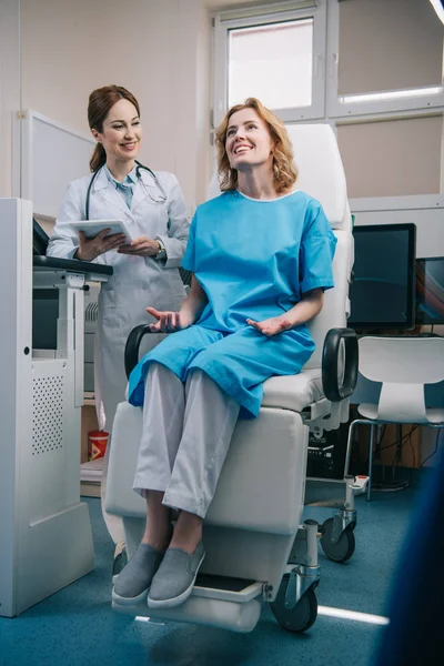 Messa a fuoco selettiva di donna felice seduta in poltrona vicino sorridente medico in possesso di tablet digitale — Foto stock