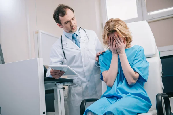 Mujer llorando sentada en un sillón cerca del médico sosteniendo la tableta digital - foto de stock