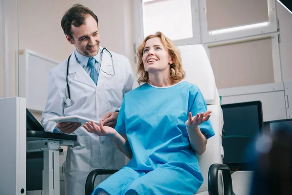 Selektiver Fokus einer glücklichen Frau, die gestikuliert, während sie im Sessel neben dem Arzt sitzt und ein digitales Tablet in der Hand hält — Stockfoto