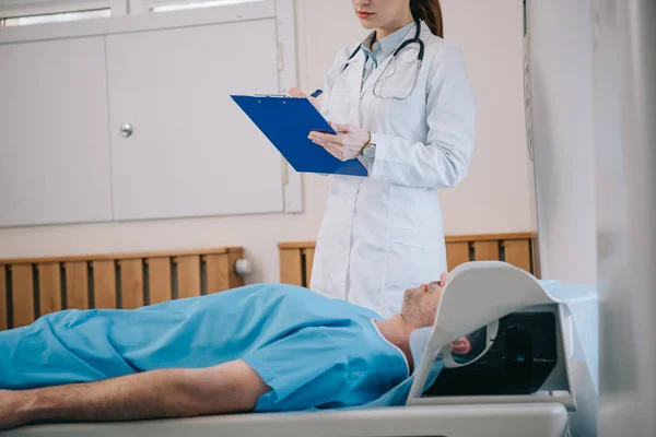 Ausgeschnittene Ansicht eines jungen Radiologen, der auf Klemmbrett schreibt, während er in der Nähe eines Patienten auf einem Mri-Scanner-Bett liegt — Stockfoto