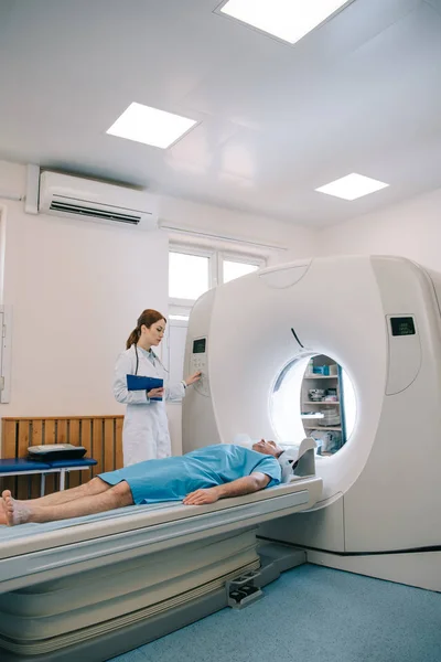 Radiologe im weißen Kittel operiert CT-Scanner, während Patient auf CT-Bett liegt — Stockfoto