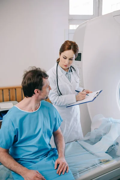 Atento médico escribiendo en el portapapeles mientras está de pie cerca del paciente sentado en la cama del escáner ct - foto de stock