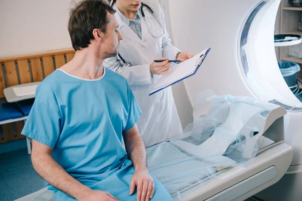 Visão parcial do jovem radiologista mostrando diagnóstico ao paciente sentado no leito do tomógrafo — Fotografia de Stock