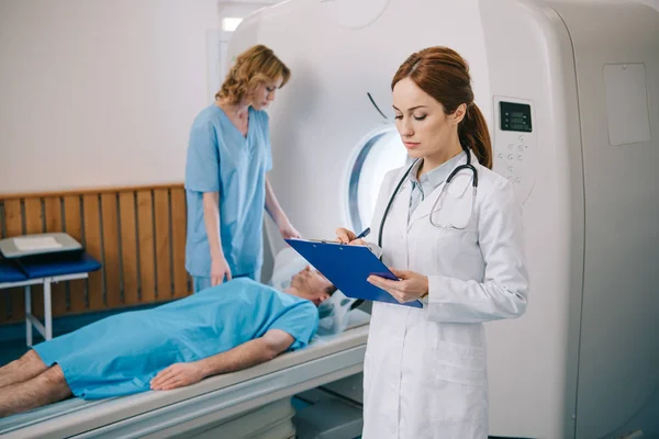 Enfermera preparando al paciente para escaneo de resonancia magnética mientras el radiólogo escribe en el portapapeles - foto de stock