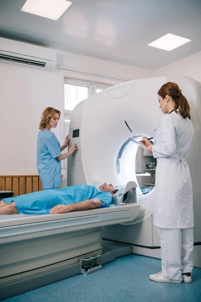 Радіолог, який пише в буфері обміну даними, коли помічник операційного сканера Кет біля пацієнта лежить на ліжку сканера — стокове фото