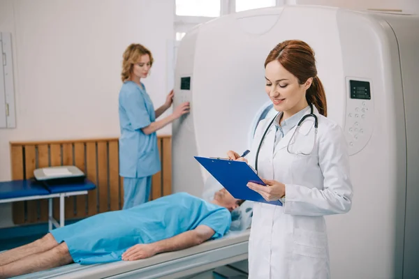 Selektiver Fokus des lächelnden Radiologen, der auf Klemmbrett schreibt, während der Assistent den ct-Scanner in der Nähe des Patienten bedient, der auf dem ct-Scanner-Bett liegt — Stockfoto