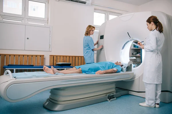 Médico escrevendo na área de transferência, enquanto em pé pela máquina mri perto assistente e paciente deitado — Fotografia de Stock