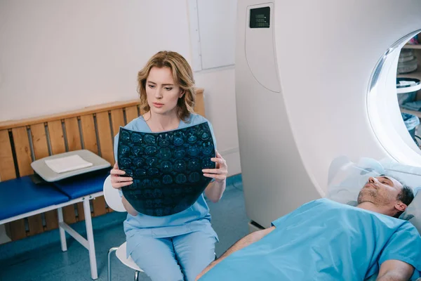 Aufmerksame Radiologin untersucht radiologische Diagnose in der Nähe des Patienten, der auf dem MRI-Bett liegt — Stockfoto