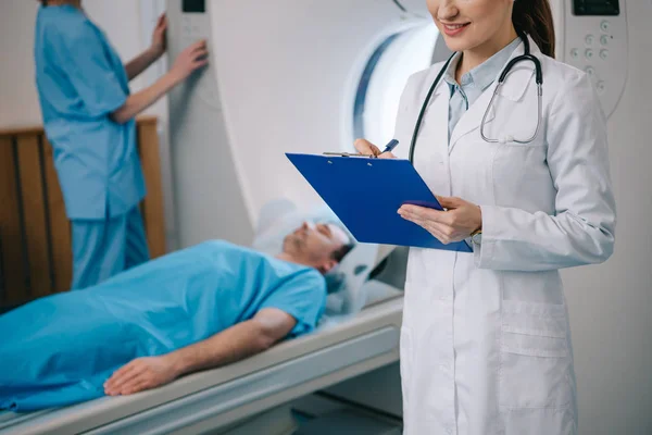 Vista recortada del radiógrafo sonriente escribiendo en el portapapeles mientras asistente de pie cerca de paciente acostado y el escáner de ct operativo - foto de stock