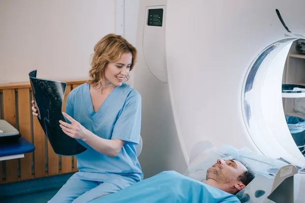 Hermoso médico sonriente con diagnóstico de radiología y mirando al hombre acostado en la cama del escáner ct - foto de stock