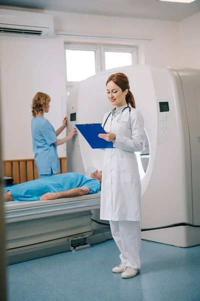 Radiografista sorrindo escrevendo na área de transferência enquanto assistente de pé perto de paciente deitado e operando scanner ct — Fotografia de Stock