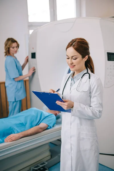 Selektiver Fokus des Radiologen, der auf Klemmbrett schreibt, während der Assistent die Maschine in der Nähe des Patienten bedient — Stockfoto