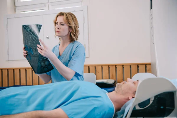 Médico atencioso examinando o diagnóstico de raios-x perto do paciente deitado no leito do tomógrafo — Fotografia de Stock