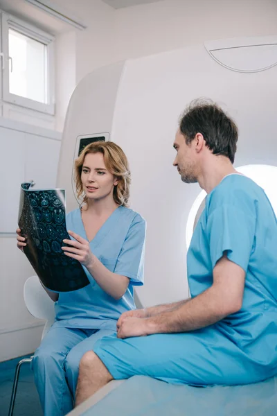 Belo médico olhando para o diagnóstico de raios-x perto do paciente sentado na cama scanner mri — Fotografia de Stock