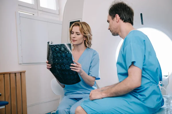 Внимательный рентгенолог, изучающий рентгеновский диагноз рядом с пациентом, сидящим на кровати КТ — стоковое фото