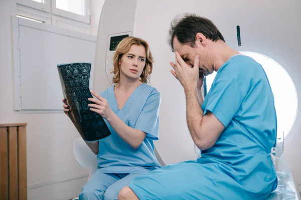 Расстроен пациент покрывая лицо руками, сидя рядом с врачом проведение рентгеновского диагноза — стоковое фото
